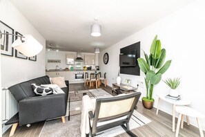 A modern living room with a sofa, a dining table with chairs, a kitchen in the background, and a large green plant. - Stylish 1-bed flat | Free parking | Fast Wi-Fi (Basingstoke)