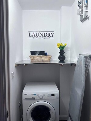 A laundry area with a washing machine, a shelf with a basket and a vase of flowers, and a wall-mounted faucet. - Stylish 1-bed flat | Free parking | Fast Wi-Fi (Basingstoke)