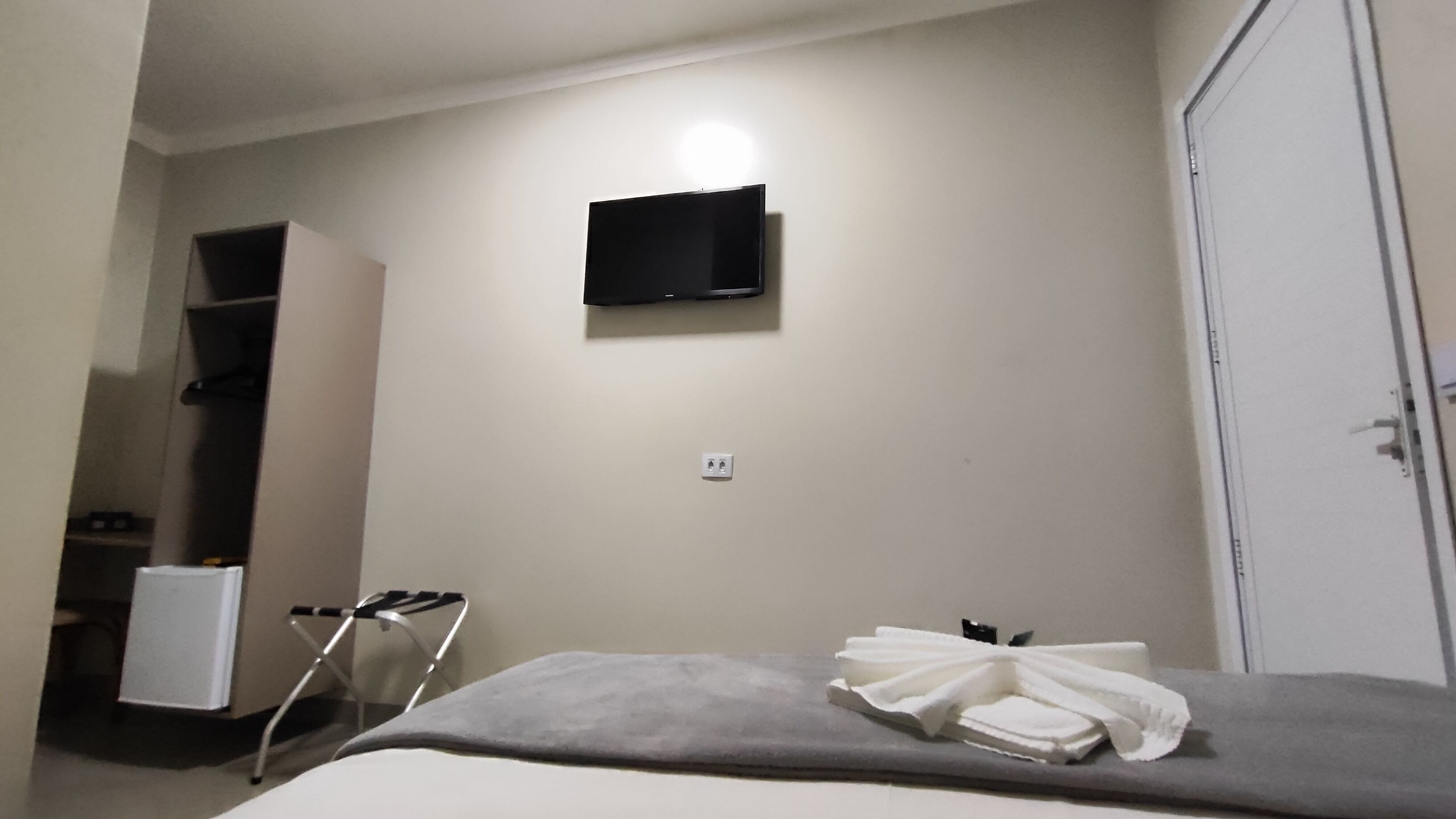A hotel room with a bed, a television mounted on the wall, a wardrobe, and a small table with a towel.