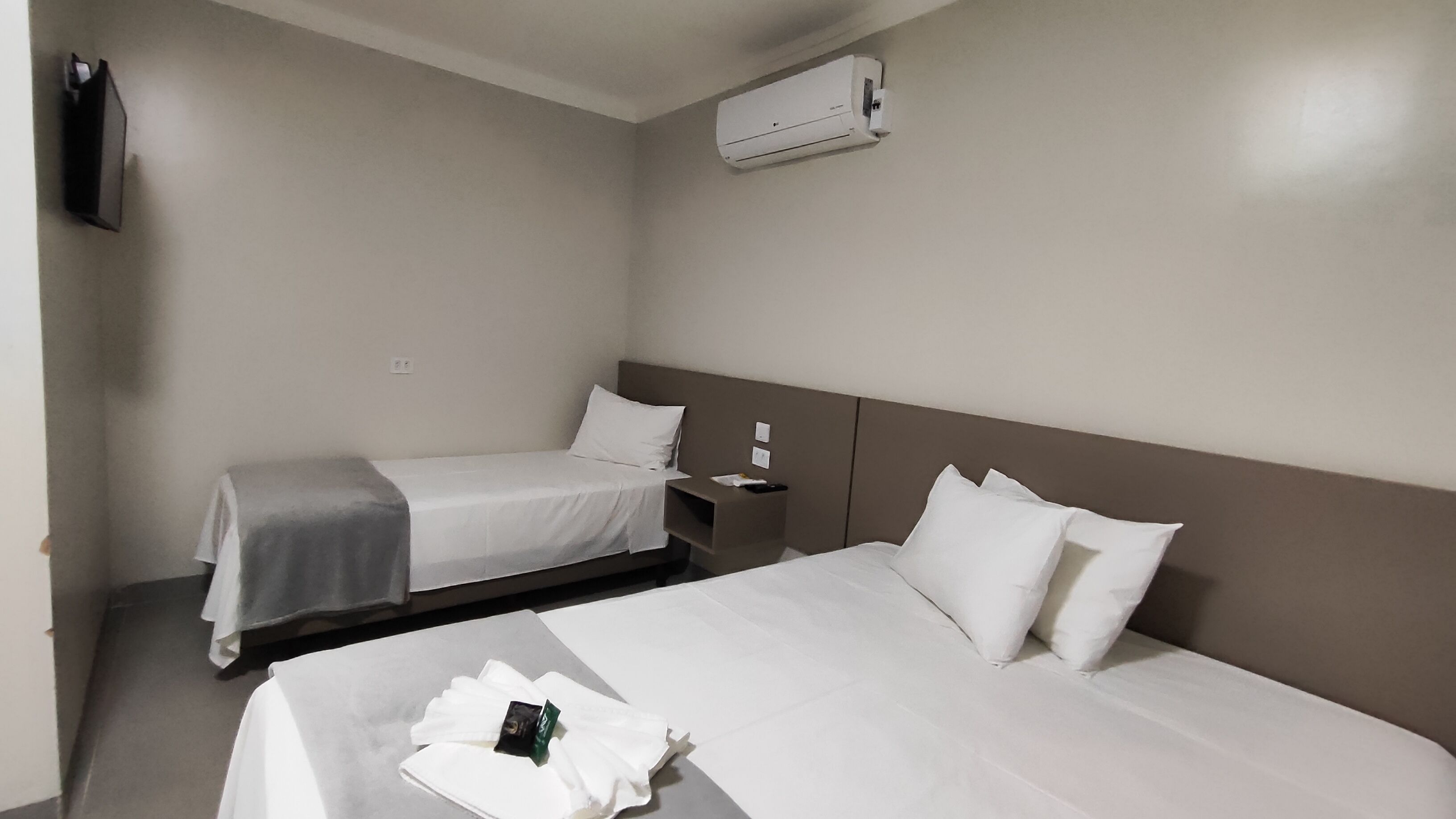 A hotel room with two beds, a TV, an air conditioner, and a bedside table with a remote control.
