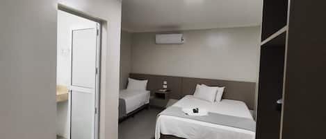 A hotel room with two beds, a bedside table, a nightstand, a wall-mounted air conditioner, and a door leading to another room.