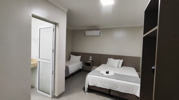 A hotel room with two beds, a bedside table, a nightstand, a wall-mounted air conditioner, and a door leading to another room.
