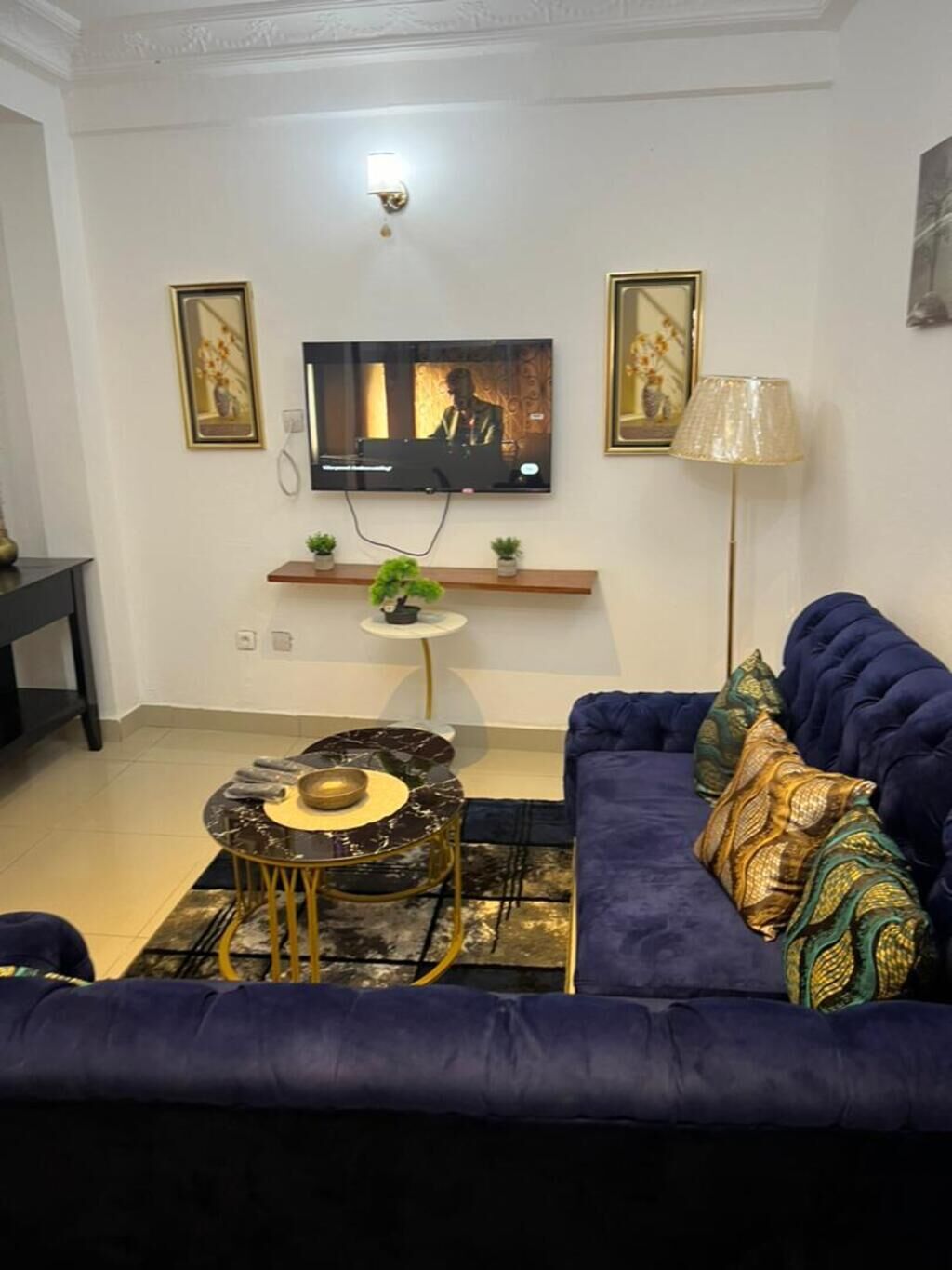 A living room with a dark blue sofa, a small round coffee table, a flat-screen TV mounted on the wall, and a floor lamp.
