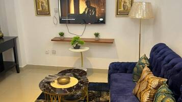 A living room with a dark blue sofa, a small round coffee table, a flat-screen TV mounted on the wall, and a floor lamp.