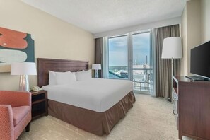 Ein Hotelzimmer mit einem großen Bett, einem Schreibtisch mit Lampe, einem Fernseher und Blick auf die Stadt durch große Fenster.