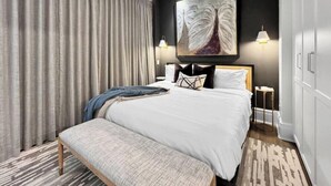 A hotel room with a large bed, a bench, a painting on the wall, and two bedside lamps. - DeLuna Vista the Brent Lofts (Pensacola)