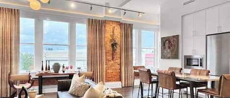 A modern living room with a sofa, a dining table with chairs, and a kitchen area with a refrigerator and microwave.