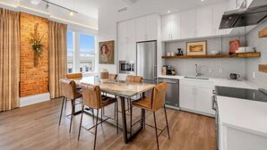 A modern kitchen with white cabinets, a stainless steel refrigerator, and a dining area with a wooden table and leather chairs. - DeLuna Vista the Brent Lofts (Pensacola)