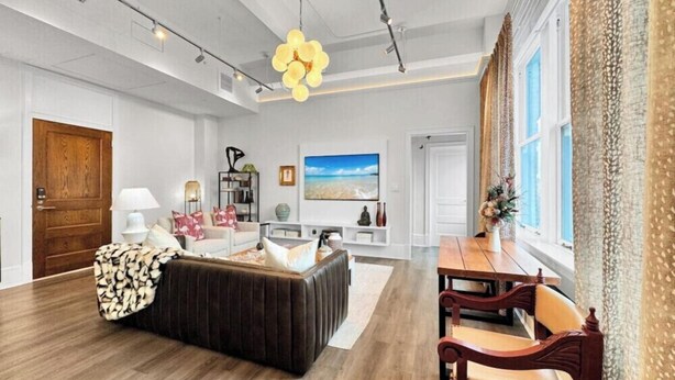 A spacious living room with a dark brown sofa, a wooden dining table with chairs, and a large window with curtains. - DeLuna Vista the Brent Lofts (Pensacola)