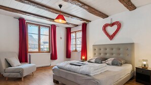 A bedroom with a bed, a chair, a nightstand, a wall-mounted heart decoration, and a window with red curtains. - Gîtes les cigogneaux - Gîte Pinot (Beblenheim)