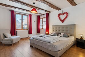 Un dormitorio con cama, una silla, una mesita de noche, una decoración con forma de corazón en la pared y una ventana con cortinas rojas.