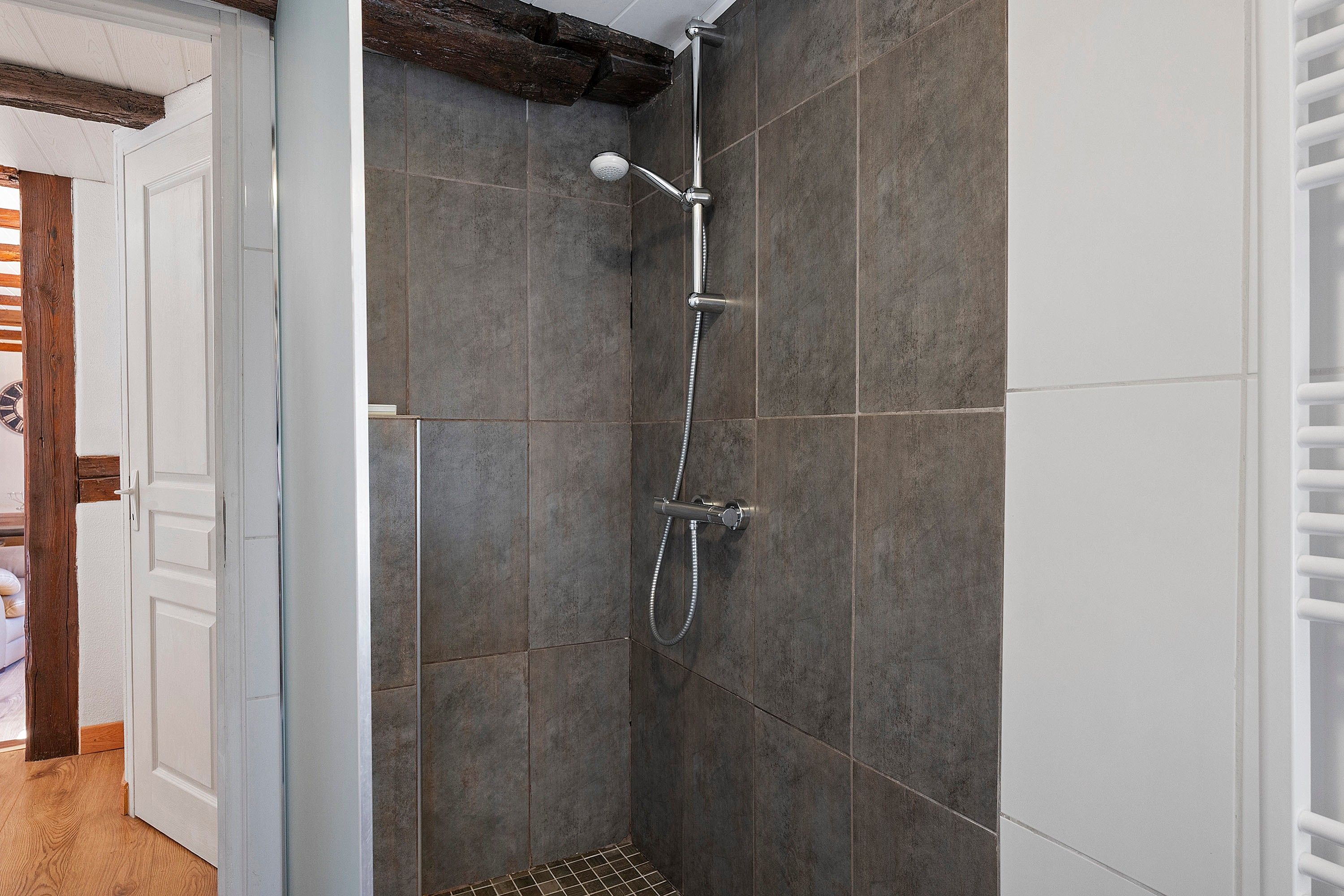 Une douche équipée d’un pommeau de douche à main et d’un robinet mural, installée dans une pièce aux murs recouverts de carreaux foncés et dotée d’une poutre en bois au plafond.