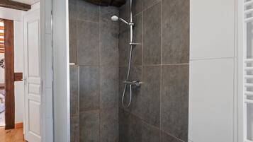 Une douche équipée d’un pommeau de douche à main et d’un robinet mural, installée dans une pièce aux murs recouverts de carreaux foncés et dotée d’une poutre en bois au plafond.