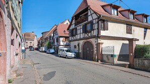 A street lined with traditional half-timbered houses, a white car parked on the side, and a clear blue sky. - Gîtes les cigogneaux - Gîte Pinot (Beblenheim)