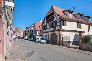 Una calle bordeada de casas tradicionales con entramado de madera, un coche blanco estacionado al lado, y un cielo azul despejado.