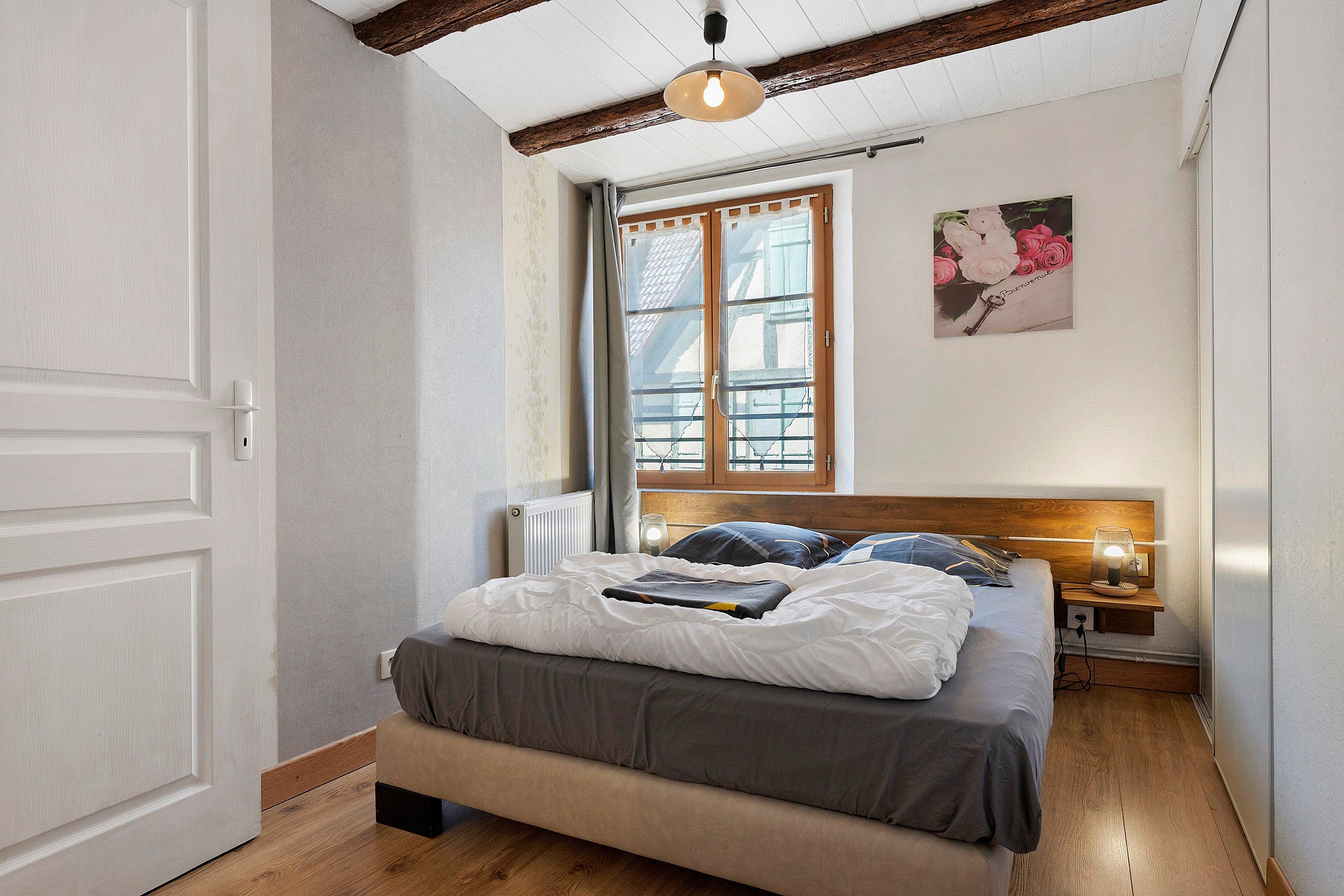 Quarto com cama, mesa de cabeceira, luminária na parede, janela com cortinas e porta.