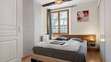 Quarto com cama, mesa de cabeceira, luminária na parede, janela com cortinas e porta.