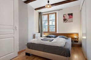 Une chambre à coucher comprenant un lit, une table de chevet, une lampe fixée au mur, une fenêtre avec des rideaux et une porte.