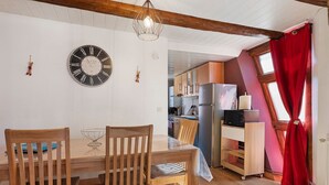 A dining area with a wooden table and chairs, a wall clock, and a kitchen visible through an open door. - Gîtes les cigogneaux - Gîte Pinot (Beblenheim)