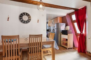 Un área de comedor con una mesa y sillas de madera, un reloj de pared y una cocina visible a través de una puerta abierta.