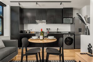 A modern kitchen with dark cabinets, a microwave, a washing machine, and a dining area with a table and chairs. - Skyline Tokyo Studio w/ Table Mountain Views & Rooftop Pool and Lounge (Cape Town)