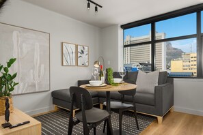 A modern living room with a dining area, a sofa, a coffee table, and a view of a cityscape through large windows. - Skyline Tokyo Studio w/ Table Mountain Views & Rooftop Pool and Lounge (Cape Town)