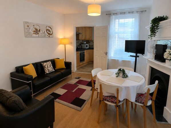 A modern living room with a black sofa, a dining table with chairs, a kitchen in the background, and a fireplace with a plant on the mantel.