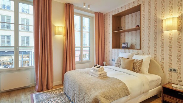 A hotel room with a large bed, a wooden headboard, a nightstand, a wall-mounted shelf, and a view of the city through the windows. - Sublim new Triplex on Place des Vosges (Paris)