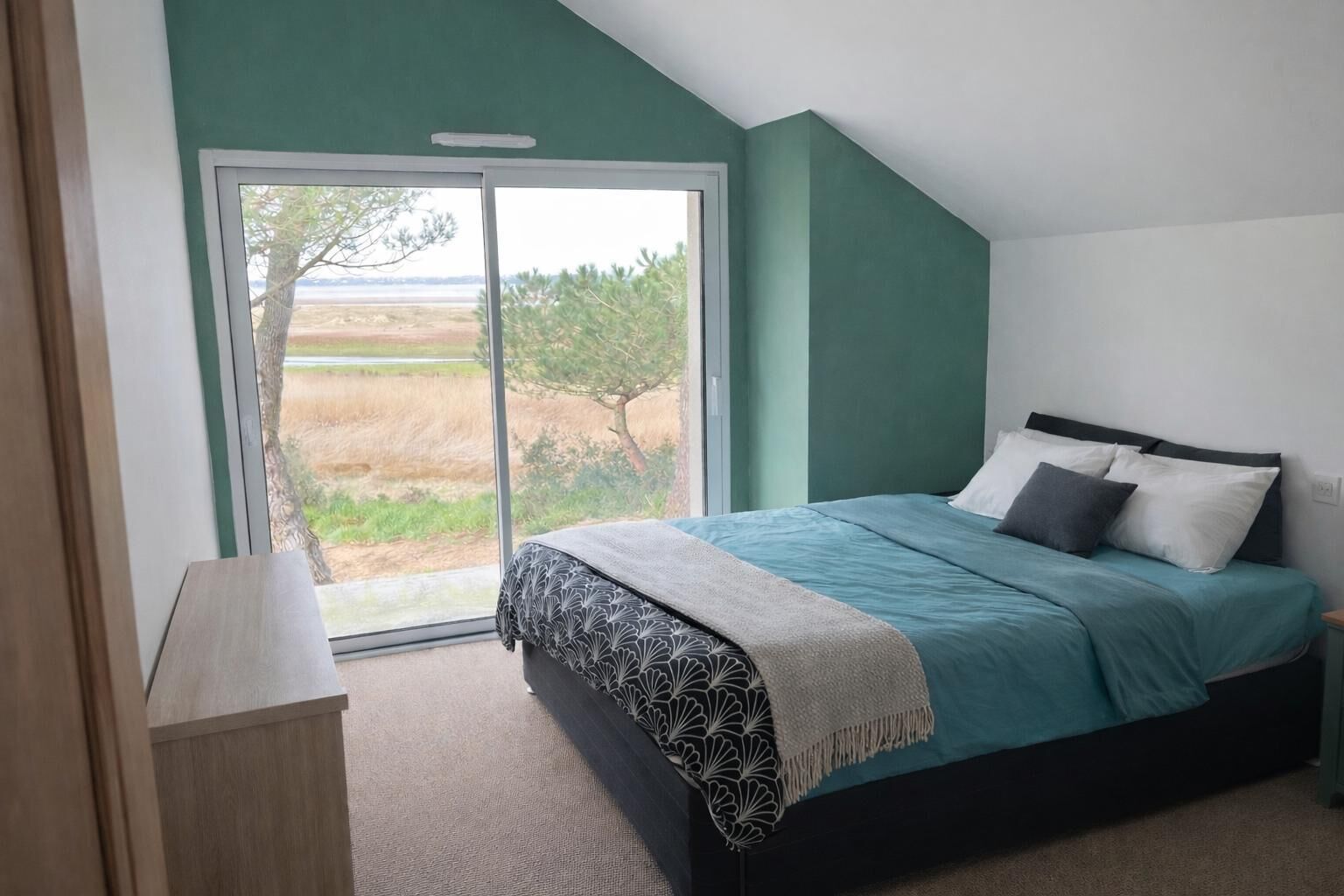 A bedroom with a bed, a nightstand, a window with a view of trees and fields, and a door.