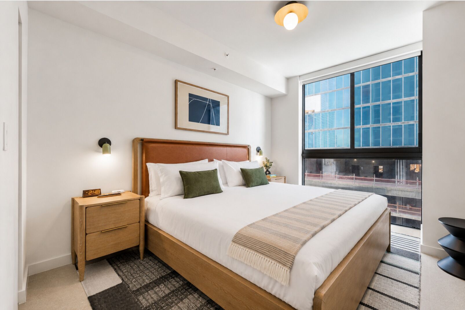 Een moderne hotelkamer met een groot bed, een nachtkastje, een wandklok, een lijst met een afbeelding en een groot raam met uitzicht.