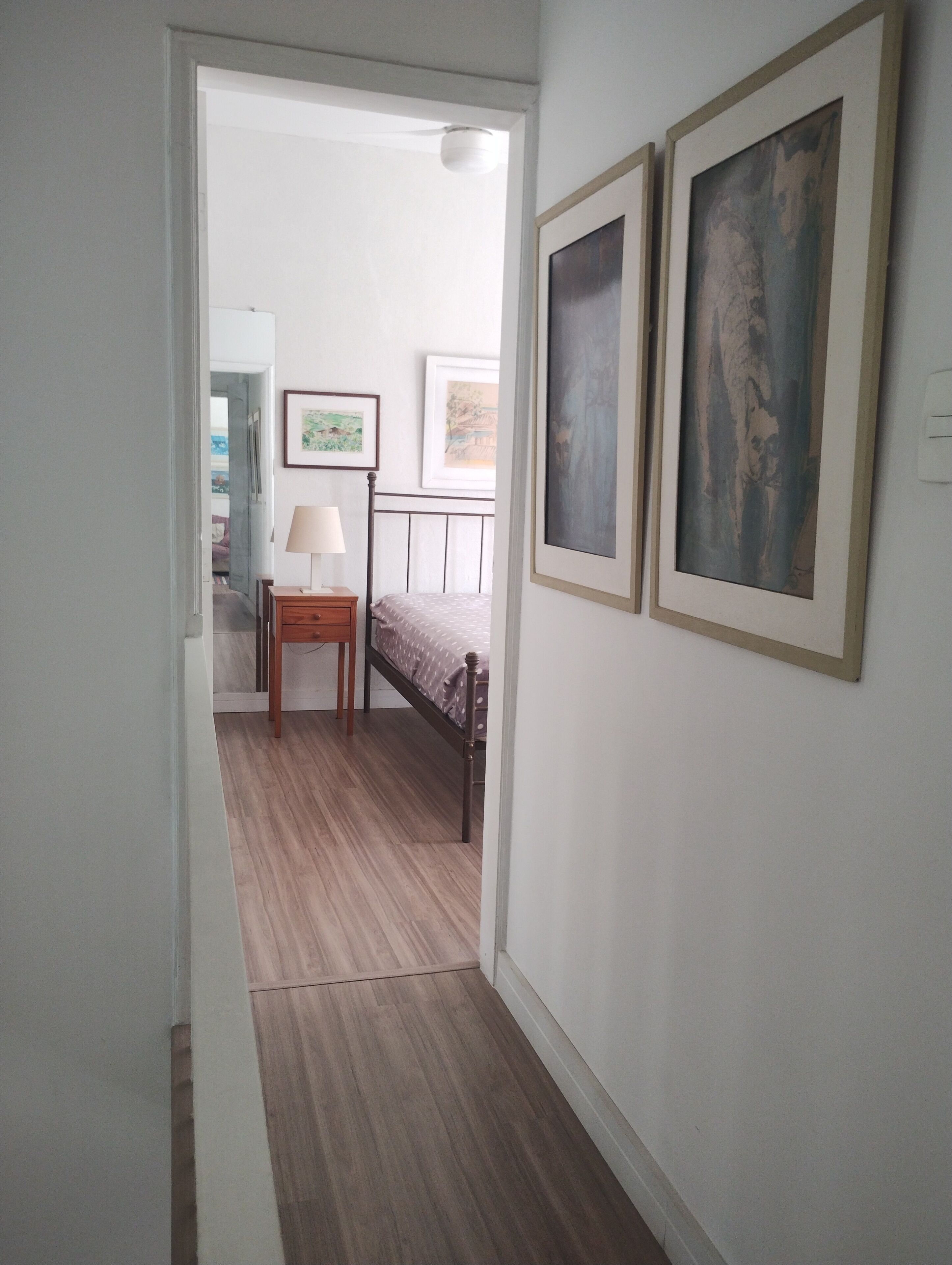 A narrow hallway leading to a bedroom with a bed, a bedside table, and a lamp. There are framed pictures on the wall.