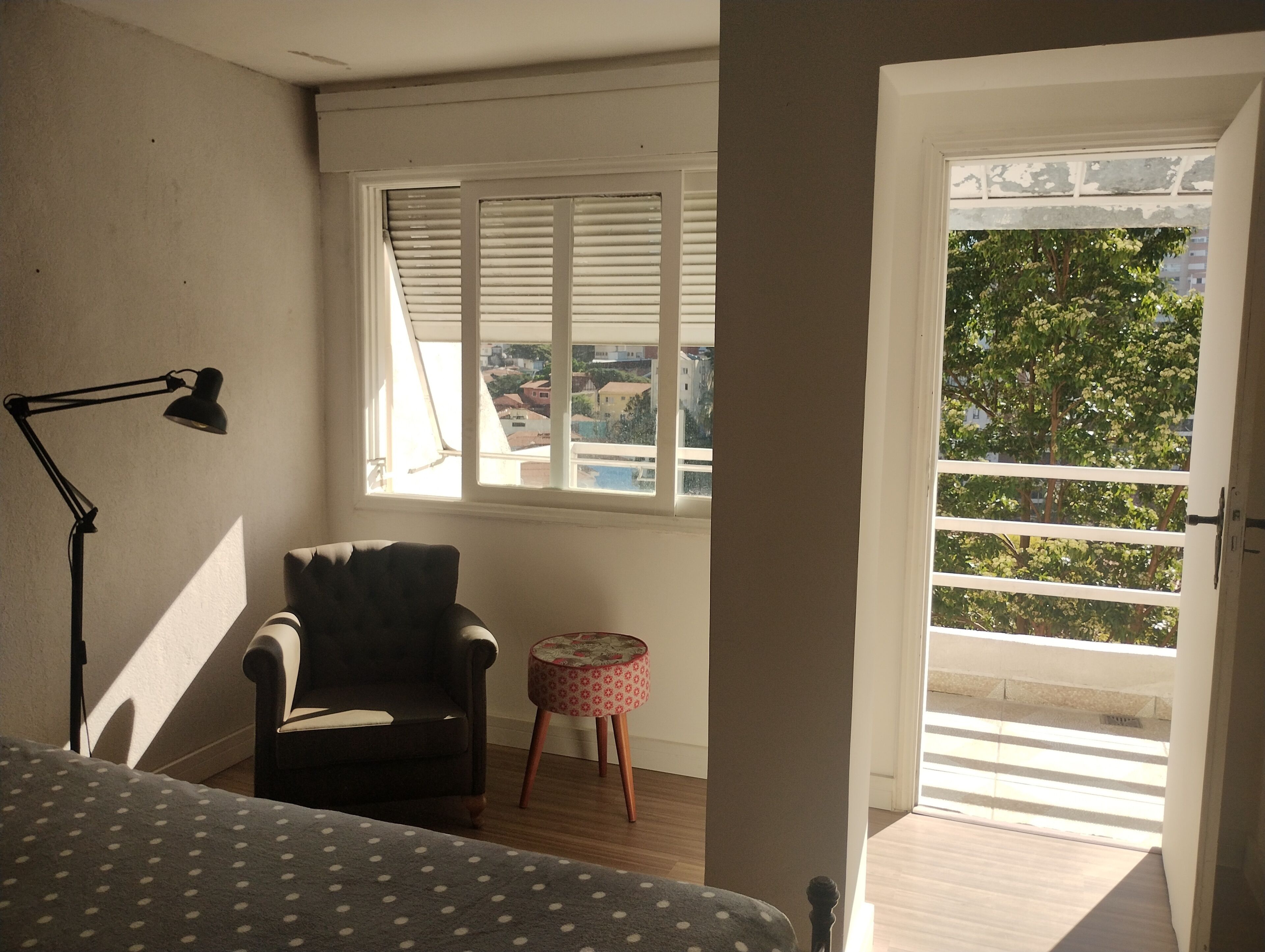 A room with a bed, a chair, a small table, a window with blinds, and a door leading outside.