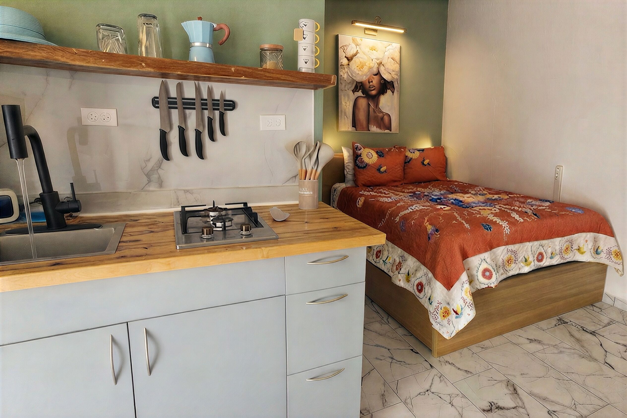 Uma cozinha compacta com fogão a gás, pia e bancada. Há uma cama com colcha floral e dois travesseiros.