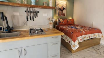 Uma cozinha compacta com fogão a gás, pia e bancada. Há uma cama com colcha floral e dois travesseiros.