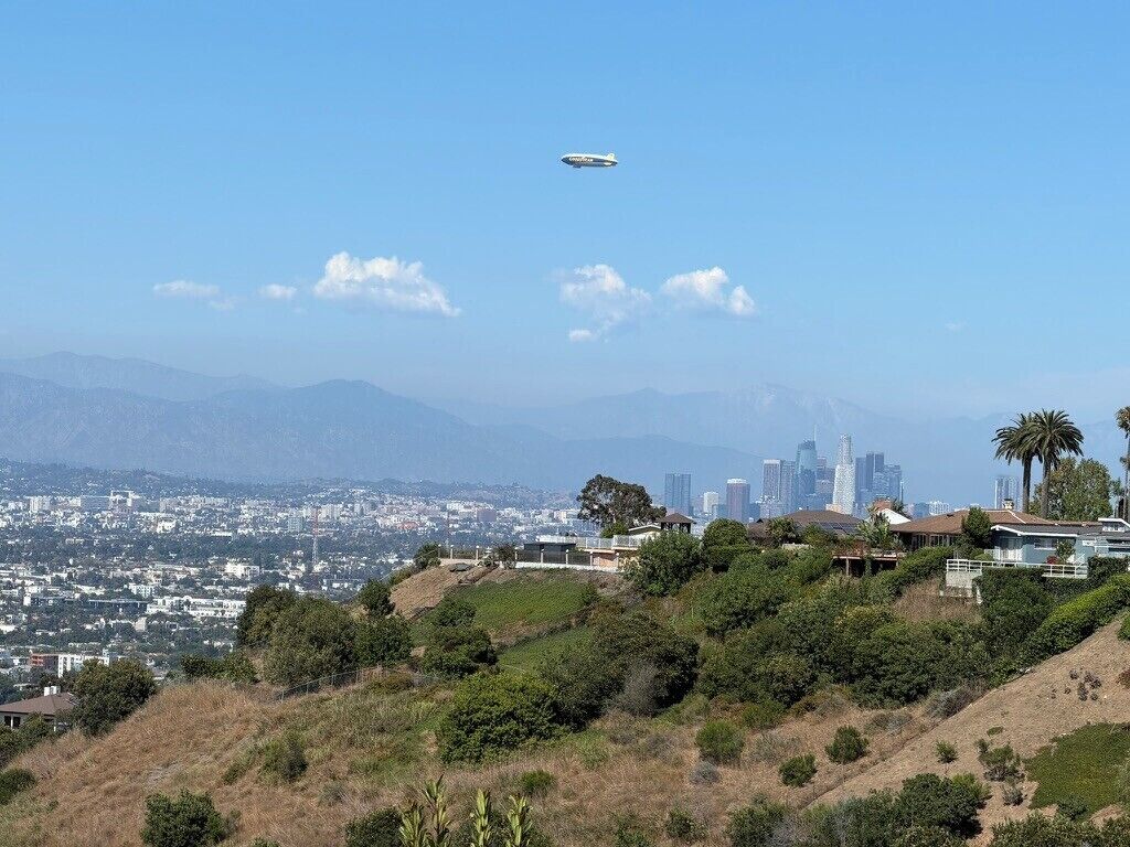 A cityscape with a clear sky, a distant mountain range, and a plane flying overhead.
