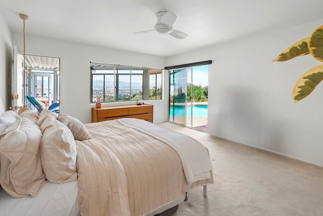 A bedroom with a bed, a dresser, a ceiling fan, and a large window with a view of a pool and cityscape.