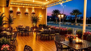 Un espacio de comedor al aire libre con mesas y sillas, plantas en macetas y vista de la calle al atardecer.