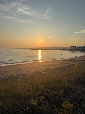 Een strand met een ondergaande zon boven het water, een paar mensen in de verte en gebouwen aan de horizon.