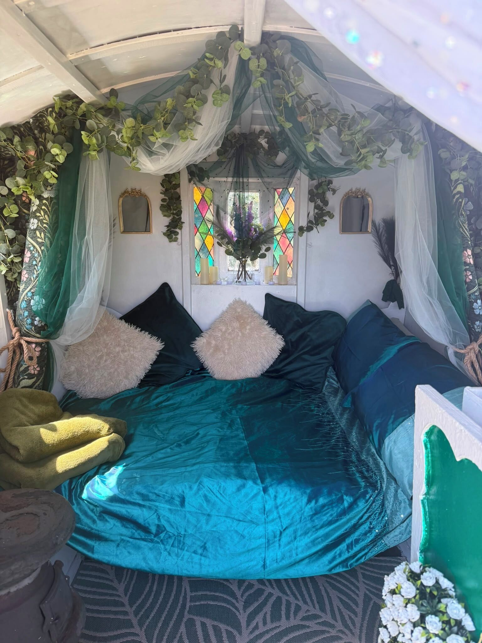 A room with a bed, pillows, a window with stained glass, and hanging plants.