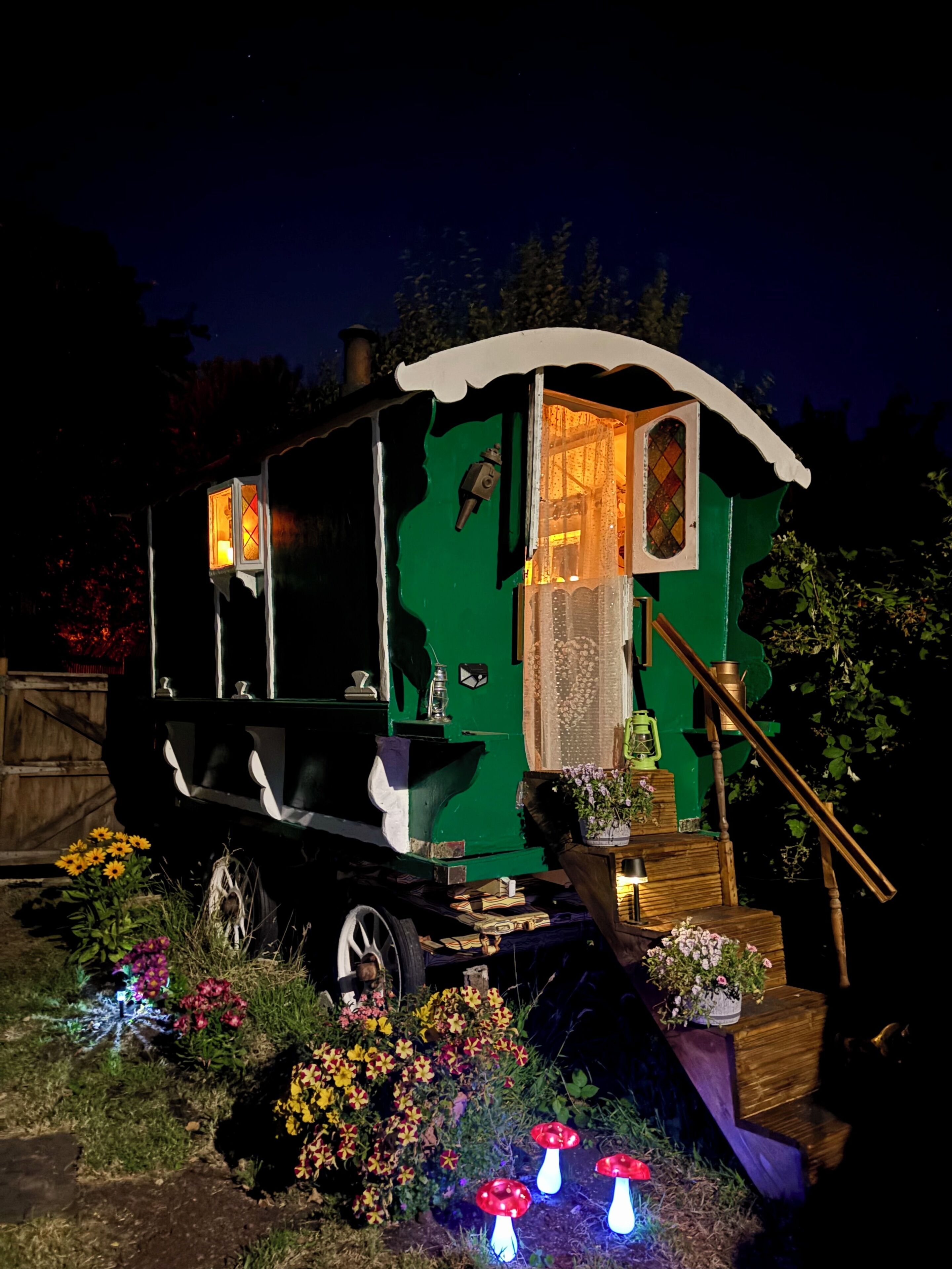 A green, vintage-style carriage with a lantern and a small window, surrounded by flowers and illuminated by garden lights.