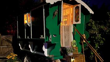 A green, vintage-style carriage with a lantern and a small window, surrounded by flowers and illuminated by garden lights.
