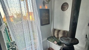 A cozy room with a visible window, a decorative curtain, a woven rug, a wall-mounted heater, and a shelf with items.