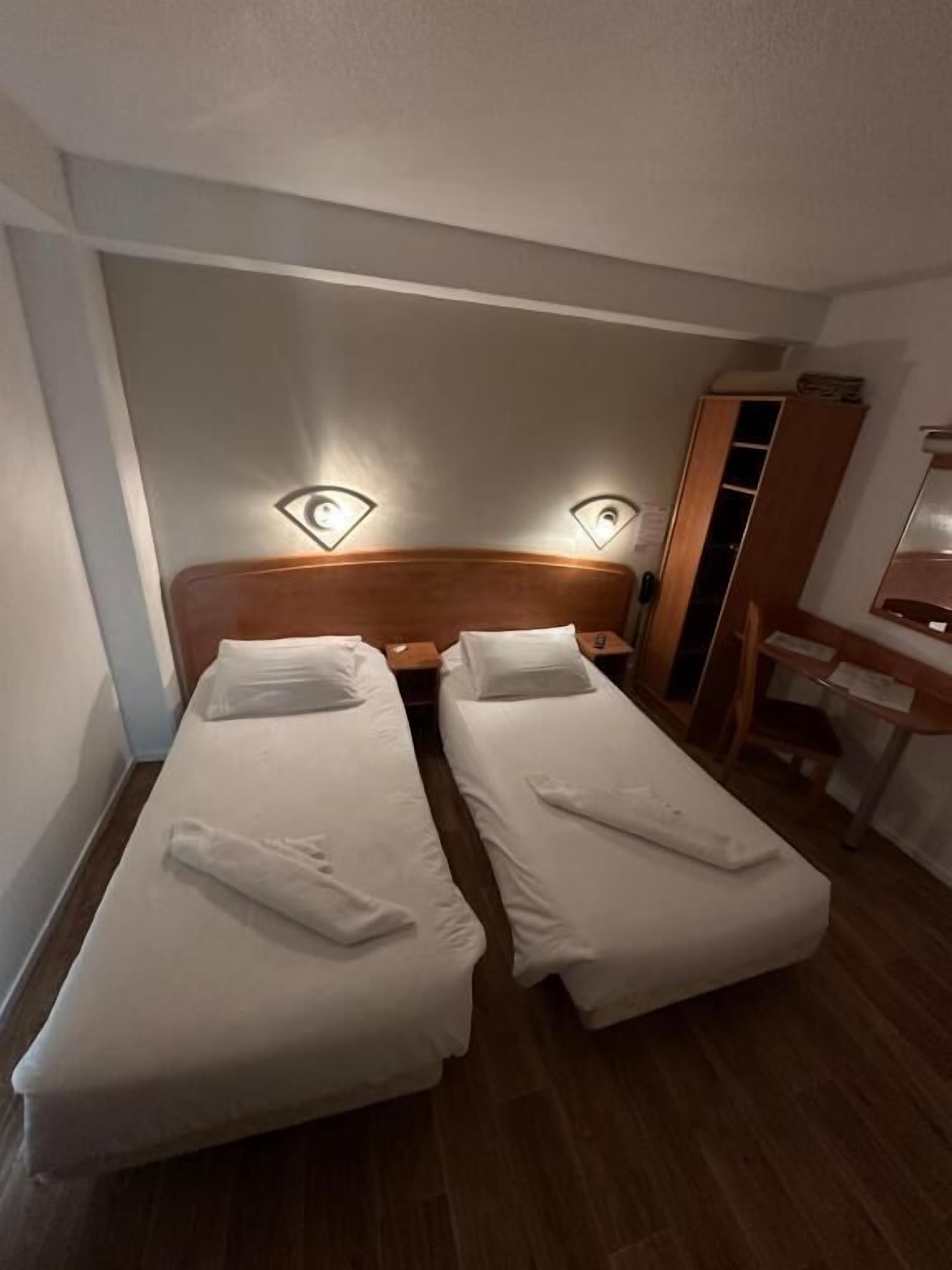 A small hotel room with two single beds, a wooden headboard, bedside lamps, a wooden wardrobe, and a desk with a chair.