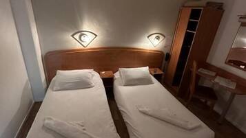 Quarto de hotel pequeno com duas camas de solteiro, cabeceira de madeira, abajures, guarda-roupa de madeira e uma escrivaninha com cadeira.