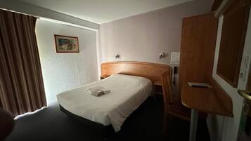 Ein Hotelzimmer mit einem Bett, einem hölzernen Kopfteil, einem Schreibtisch mit Stuhl, einem Spiegel und einem Fenster mit Vorhängen.