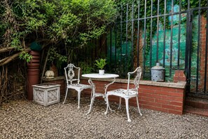 Une terrasse avec deux chaises en métal blanc et une table, entourée de verdure et d’un mur de briques.