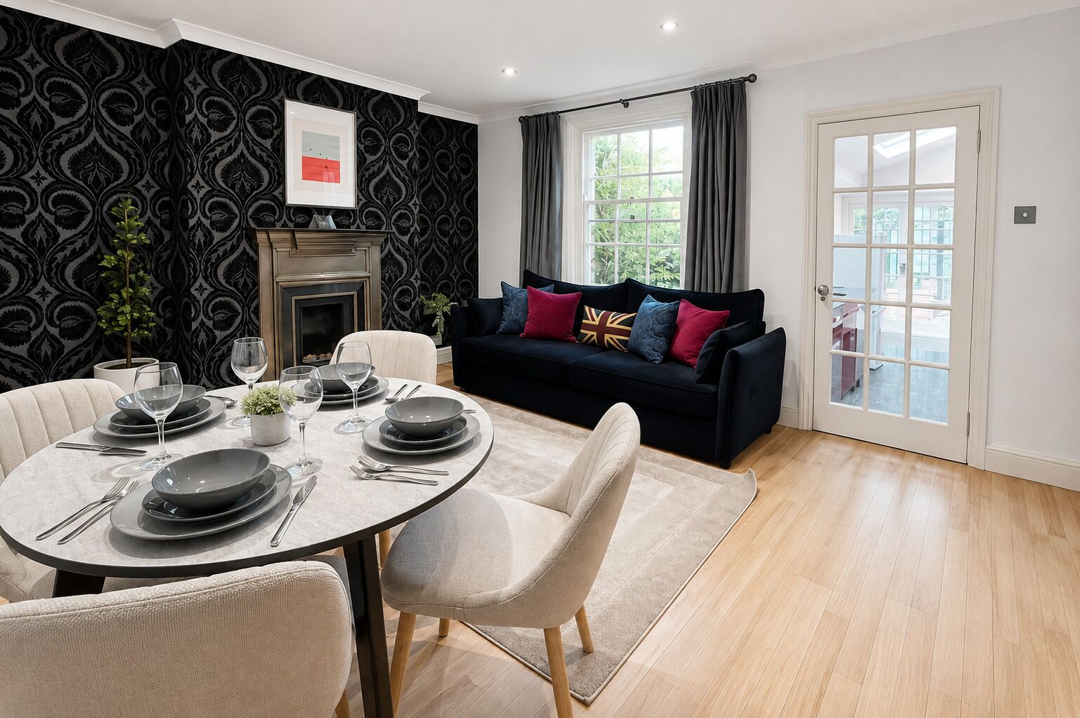 A dining area with a round table set for four, a sofa with colorful cushions, a fireplace, and a view through a window.