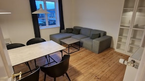 En moderne stue med trægulv, en grå sofa, et spisebord med stole og et vindue med gardiner.