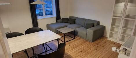 A modern living room with a wooden floor, a grey sofa, a dining table with chairs, and a window with curtains.