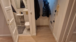En smal gang med et badeværelse til venstre, der inkluderer toilet, håndvask og spejl. Der er to døre til højre, hvoraf den ene er åben.